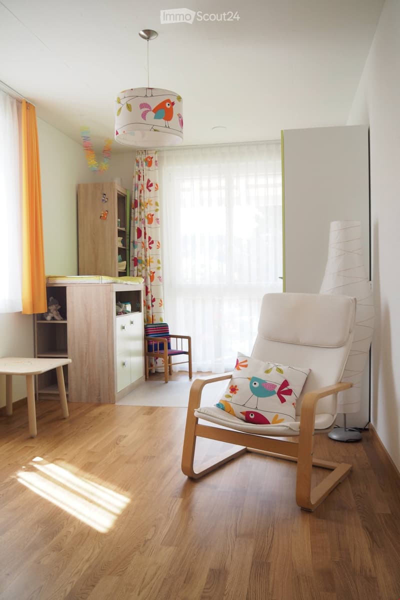 Brightly lit bedroom with wooden flooring, a white armchair with a decorative pillow, a small table, a wooden cabinet, and a hanging lamp with a bird-patterned shade.