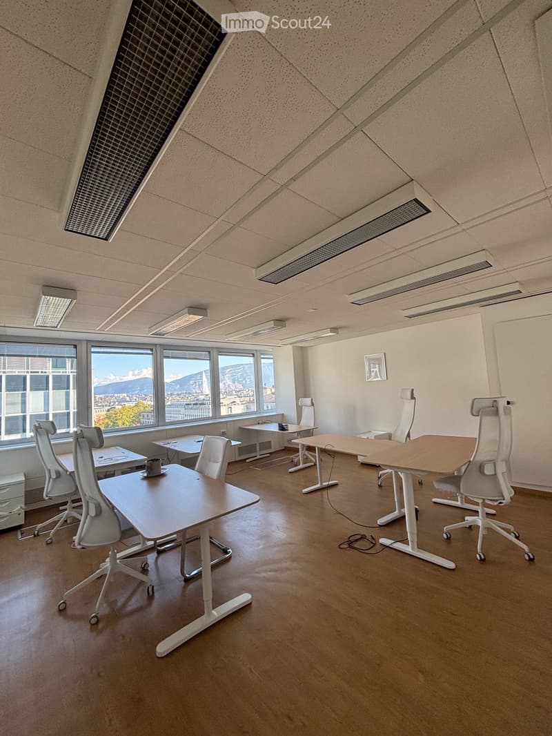 Modern office space with multiple desks, ergonomic chairs, and a cup on one of the desks. Large windows offer a scenic mountain view.