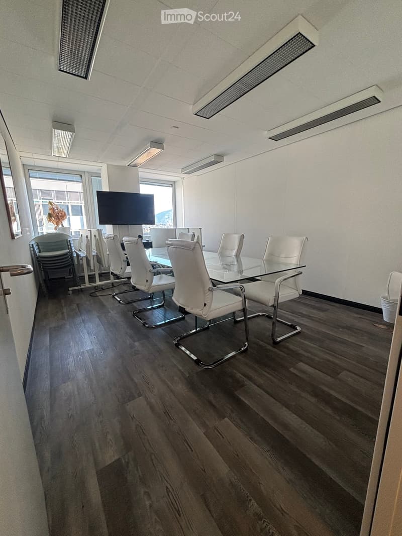 Modern conference room with wooden flooring, white walls, and large windows. Features a glass table surrounded by white chairs and a large screen mounted on the wall.