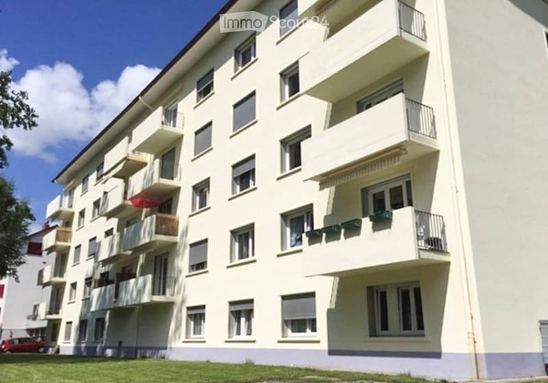 Multi-story residential building with multiple balconies and windows, light-colored facade, and a small lawn area in front.