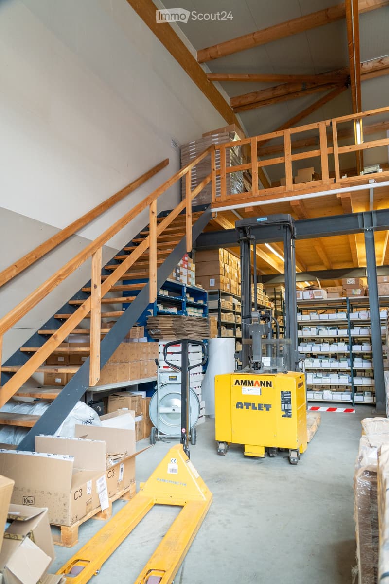 Warehouse with high ceilings, wooden beams, and industrial equipment including forklifts and pallet jacks. Shelves filled with boxes and storage items are visible, along with a wooden staircase leading to an upper level.