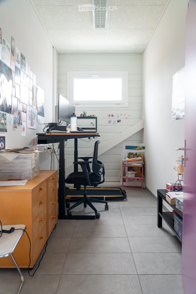A modern, well-lit room with a desk, chair, screen, and shelves. The room features a large window and a staircase leading to another level.