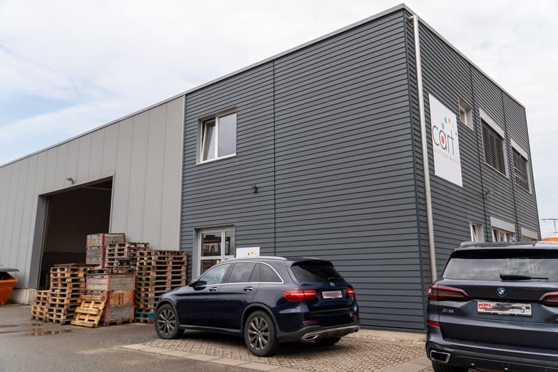 Modern industrial-style building with grey siding, large windows, and a prominent sign. Two luxury cars are parked in front, and wooden pallets are stacked near the open garage entrance.