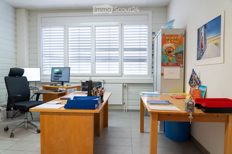 Office space with two desks, chairs, computer screens, keyboards, and various office supplies. Large windows with blinds and posters on the walls.