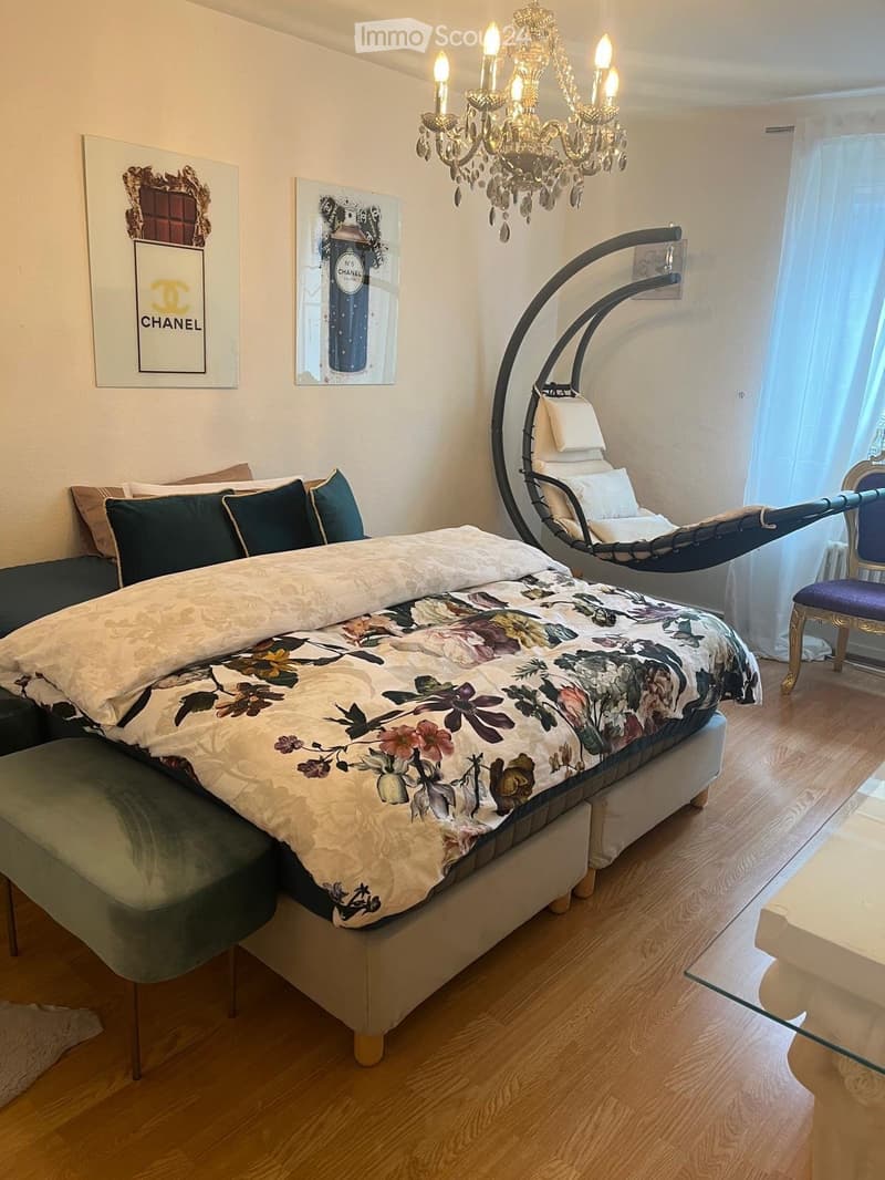 Elegant bedroom with a large bed, floral duvet, chandelier, sofa, and a hanging chair. Features wooden flooring and artwork on the walls.