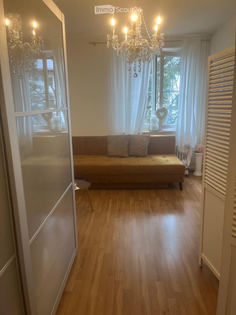A well-lit living room with wooden flooring, a beige sofa with patterned cushions, a small round table, and two chandeliers hanging from the ceiling. Large windows with white curtains allow natural light to fill the space.