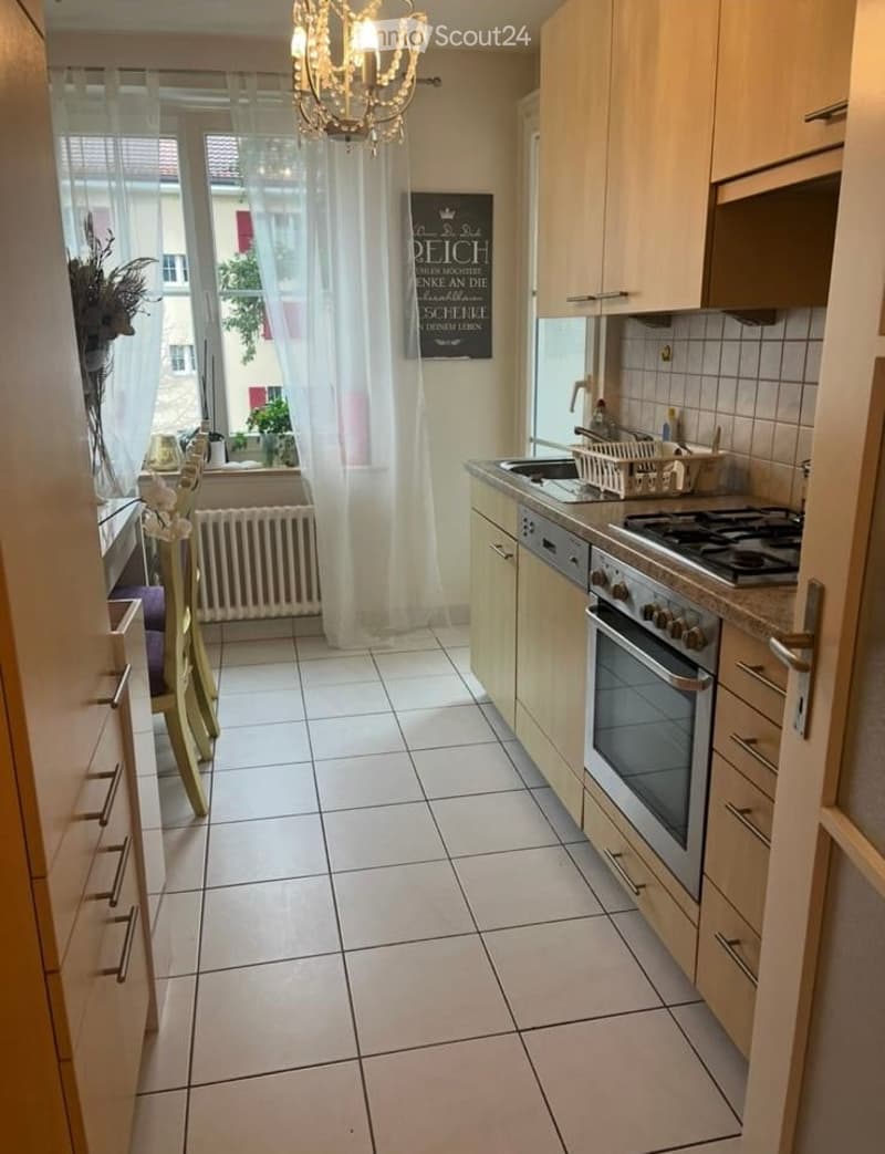 A modern kitchen with white tiled flooring, light wooden cabinets, and stainless steel appliances including an oven and sink. A chandelier hangs from the ceiling, and a window with sheer curtains lets in natural light. A small dining area with a table and
