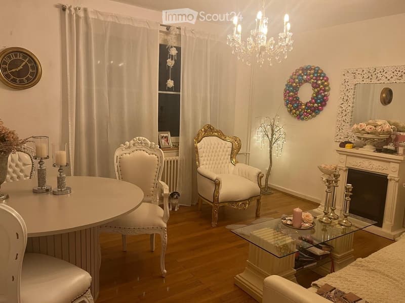 Elegant living room with white walls, wooden flooring, a round table with chairs, a white sofa, a fireplace with candles, a decorative mirror, a chandelier, and a colorful wreath on the wall.