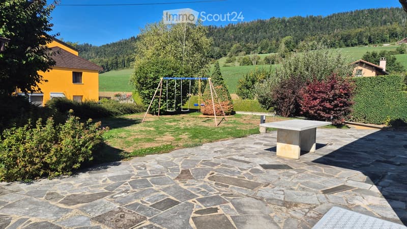 Garden area with stone-paved patio, swing set, and outdoor table surrounded by greenery and hills.