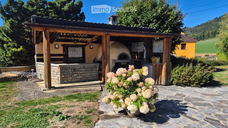 Outdoor patio with a stone pizza oven, wooden pergola, stone seating area, and surrounding greenery.