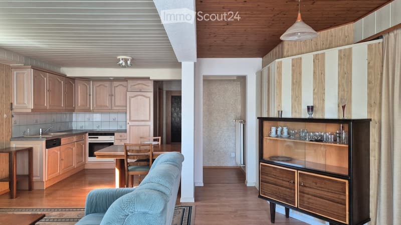 A modern, well-lit living and dining area with a sofa, dining table, chairs, cabinets, and a kitchen area with a sink, dishwasher, and oven.