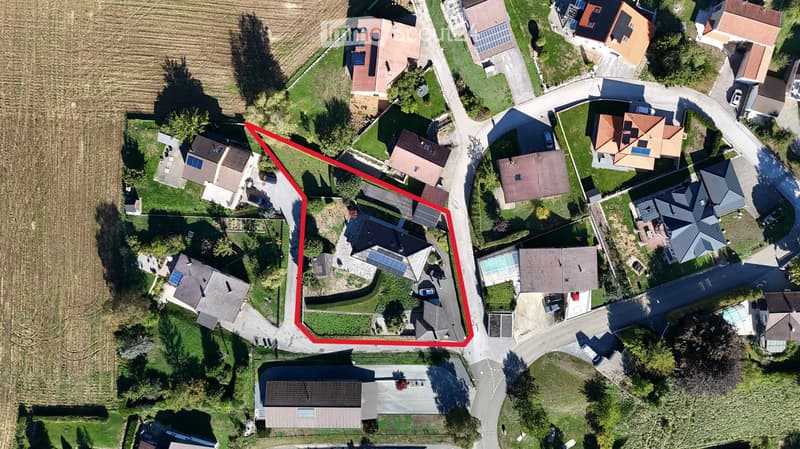 Aerial view of a residential area with multiple houses, gardens, driveways, and surrounding greenery.
