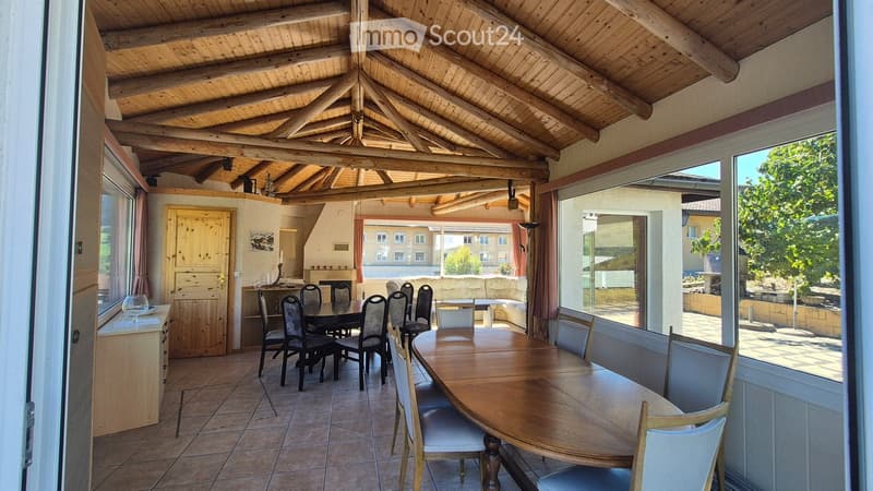 A spacious, well-lit dining area with a wooden dining table and chairs, a sofa, and a fireplace, featuring large windows and a wooden ceiling with exposed beams.