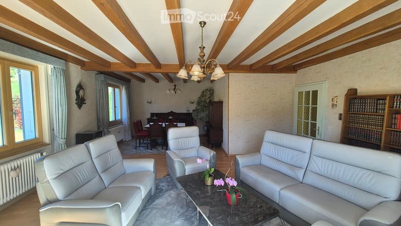Spacious living room with exposed wooden beams, white leather sofas, a coffee table with flower arrangements, a bookshelf, and a dining area visible in the background.