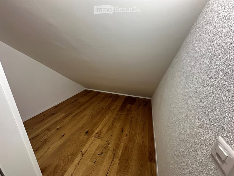 Empty room with wooden flooring and white walls, featuring a slanted ceiling and a visible light switch on the wall.