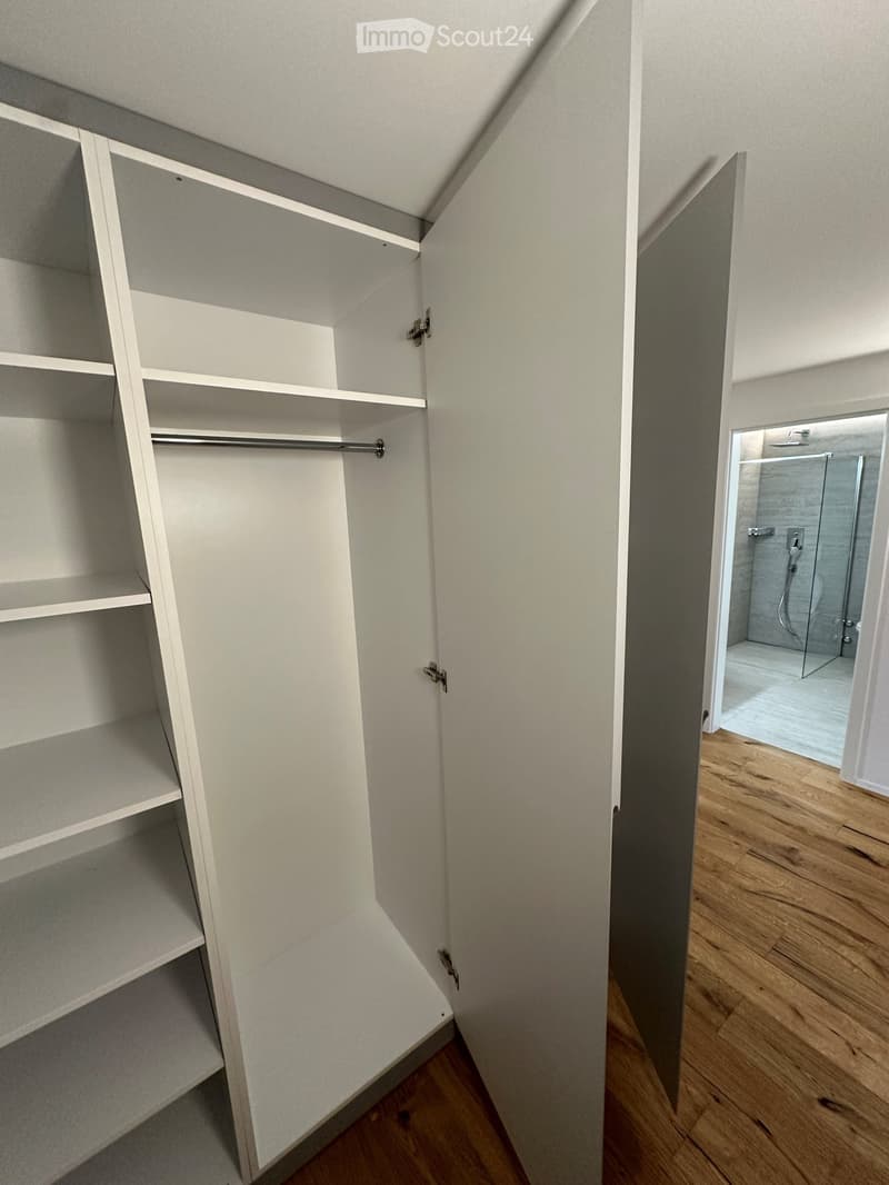 Empty closet with white shelves and a hanging rod, adjacent to a wooden-floored hallway leading to a modern shower stall with glass enclosure.