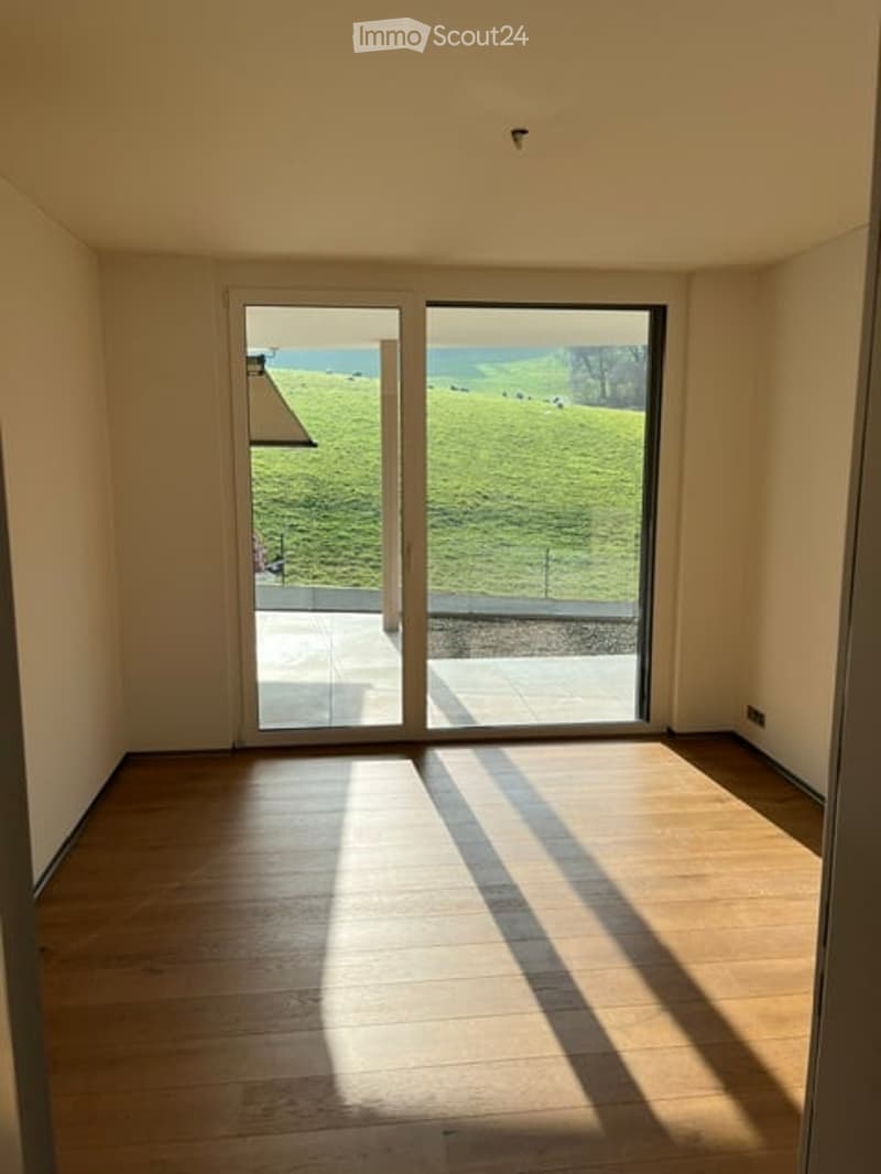 Empty room with wooden flooring, large sliding glass doors leading to a green outdoor area, and natural light streaming in.