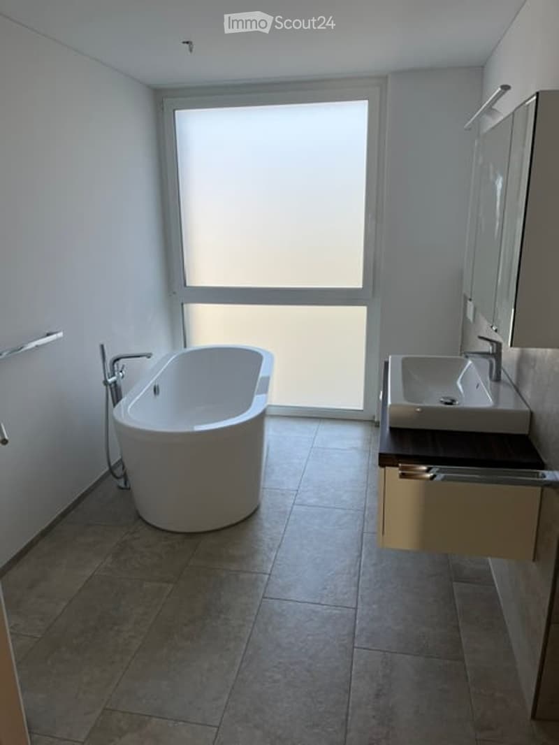 Modern bathroom with a freestanding bathtub, a sink with a mirror, and a cabinet. The space features large windows and tiled flooring.