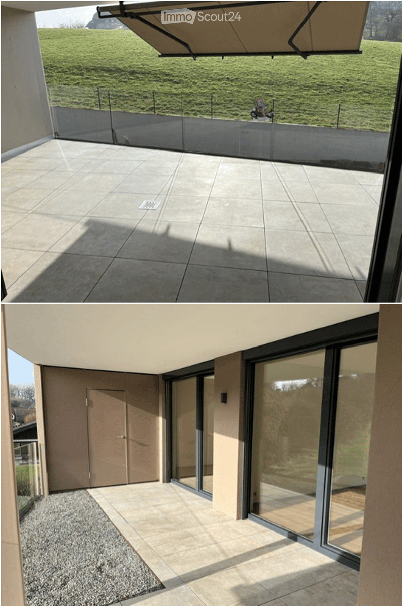 Modern balcony with large glass railing, tiled floor, and retractable awning, overlooking a green field.