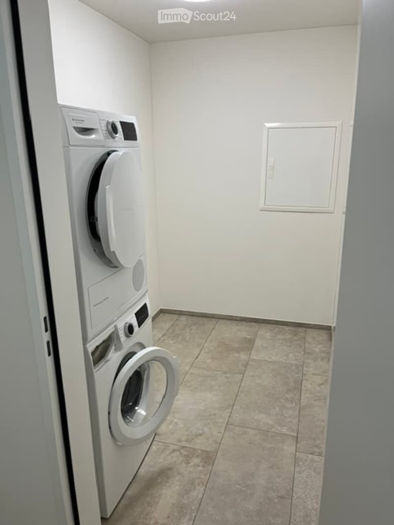 A small, clean laundry room with white tiled flooring and white walls. It features a stacked washing machine and dryer unit against one wall, with a small window above.