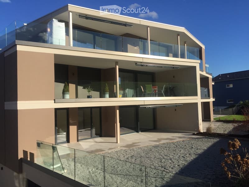 Modern two-story house with large glass balconies, beige and brown exterior, and a raised ground floor.