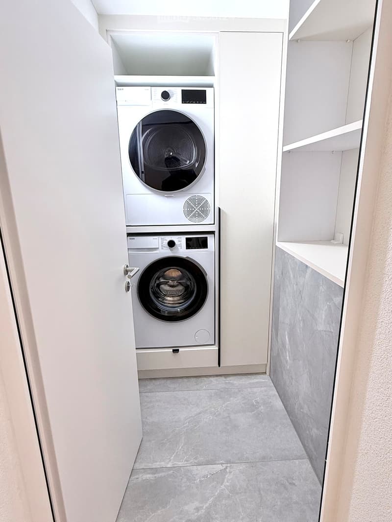 Modern laundry room with a stacked washer and dryer unit, white cabinets, and marble flooring.