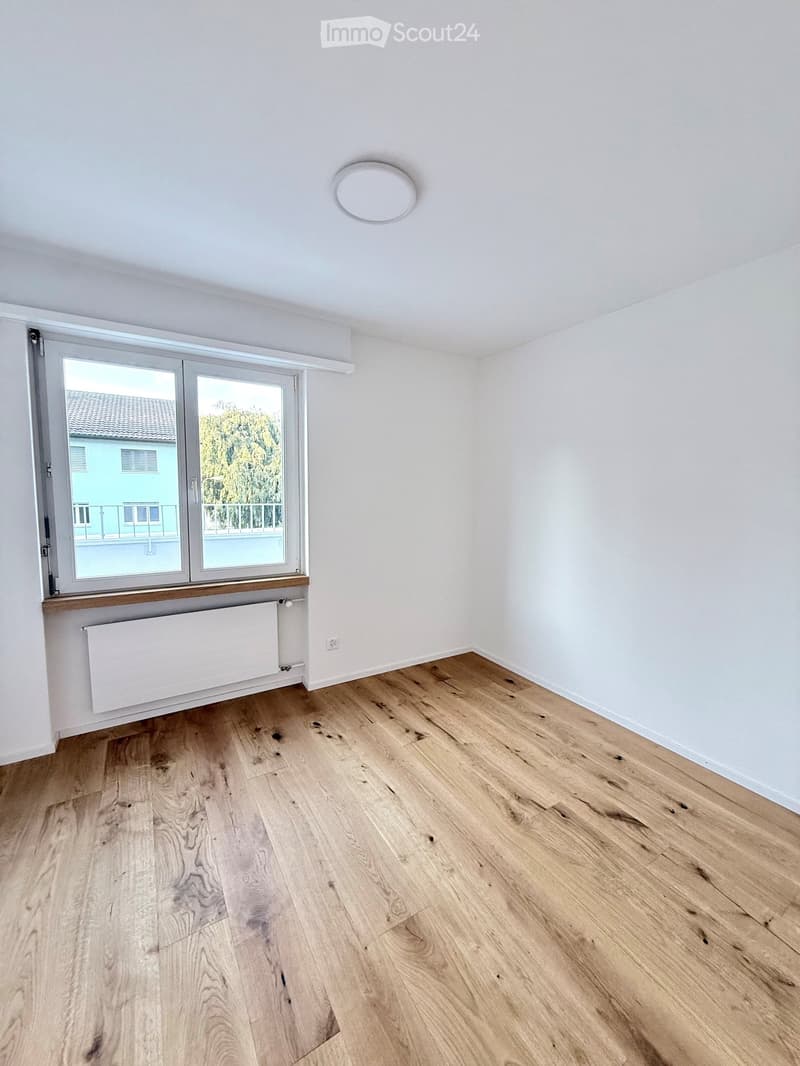 Empty room with light wooden flooring, white walls, and a large window with a view of a building and greenery outside. A radiator is visible below the window.