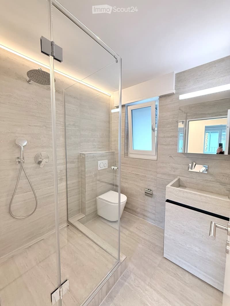 Modern bathroom with a glass-enclosed shower, toilet, sink, and mirror. Features light-colored tiles and sleek fixtures.