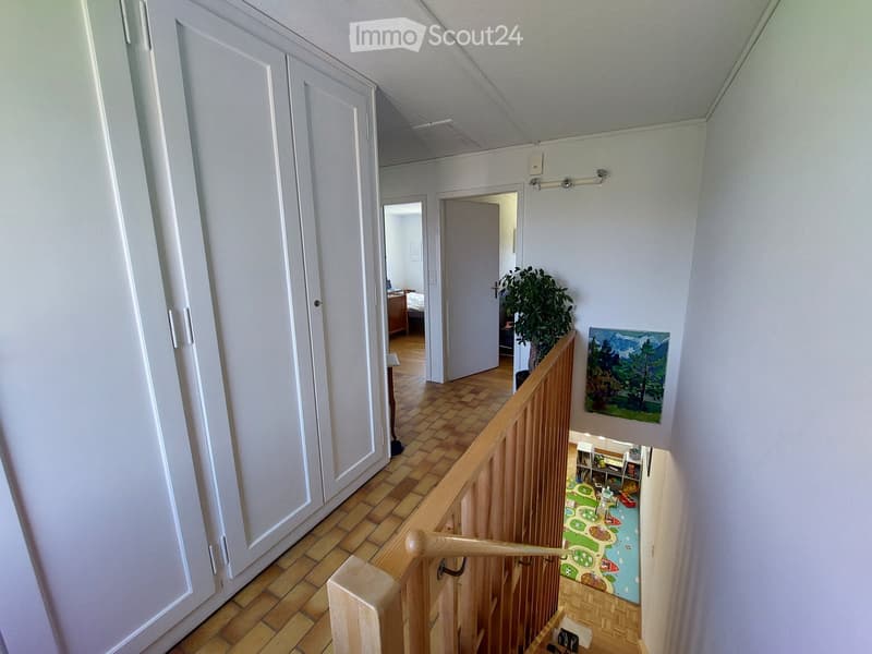 A well-lit hallway with wooden flooring, white walls, and a staircase leading to another level. The hallway features a painting on the wall, a small plant, and a glimpse of a bedroom and a child's play area in the background.