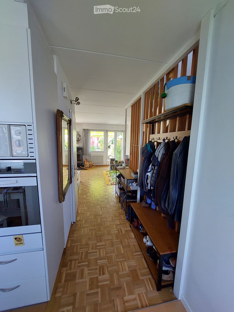 Hallway with wooden flooring, coat rack, shoe bench, and a view into a living area with a rocking chair and a small table.