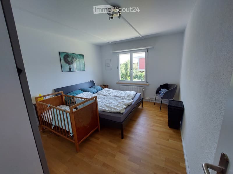Bright bedroom with wooden flooring, a double bed with pillows and blankets, a wooden baby crib, a grey armchair, a small black stool, and a picture frame on the wall.