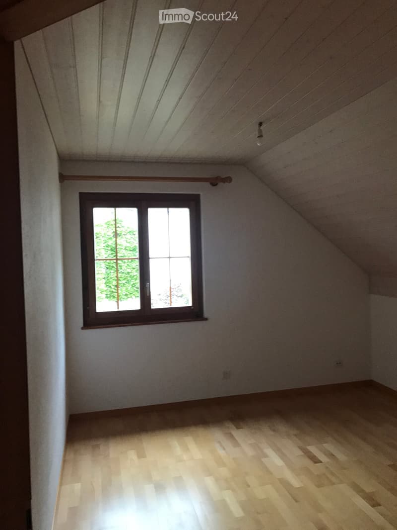 Empty attic room with wooden flooring, white walls, and a single window showing greenery outside.
