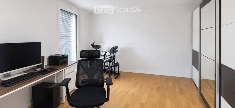A modern, well-lit room with a wooden floor, a large window, a black ergonomic chair, a desk with a computer setup, and a drum set in the corner.