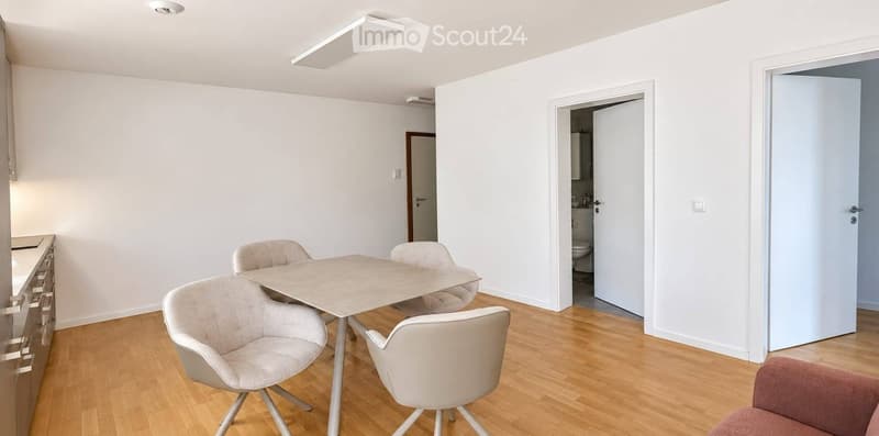 Bright, minimalist room with light wooden flooring, white walls, and modern furniture including a dining table with four chairs and a cabinet.