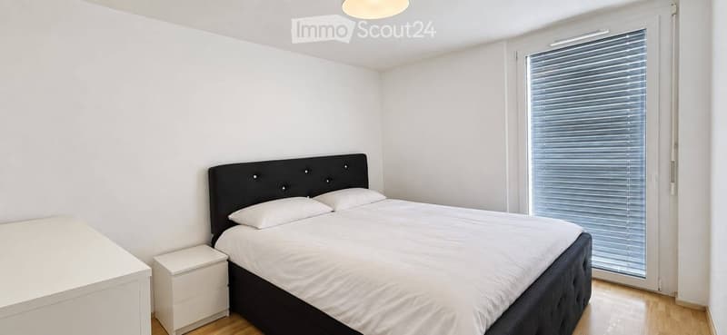 Modern bedroom with a double bed, white bedding, a nightstand, and a large window with blinds.