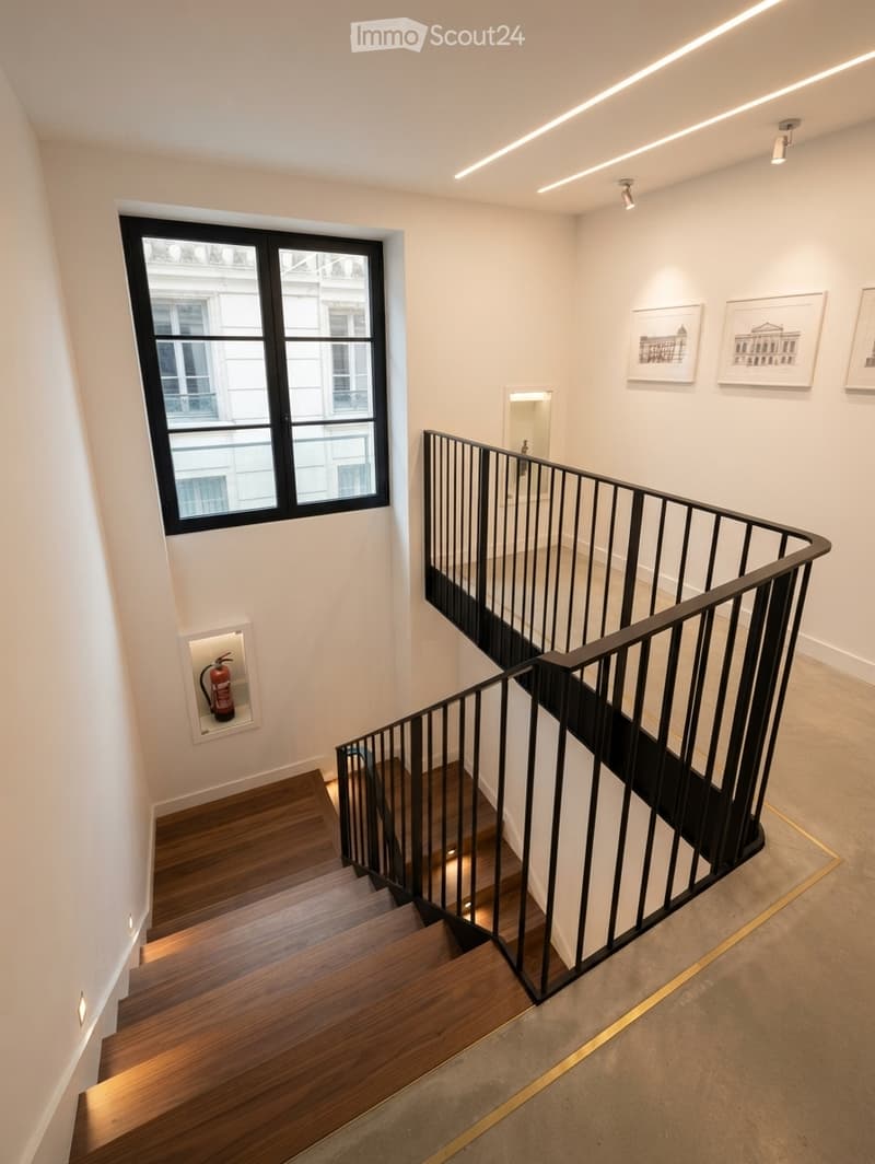 Modern staircase with wooden steps, black metal railing, and integrated lighting. Features a large window, framed artwork, and a fire extinguisher mounted on the wall.