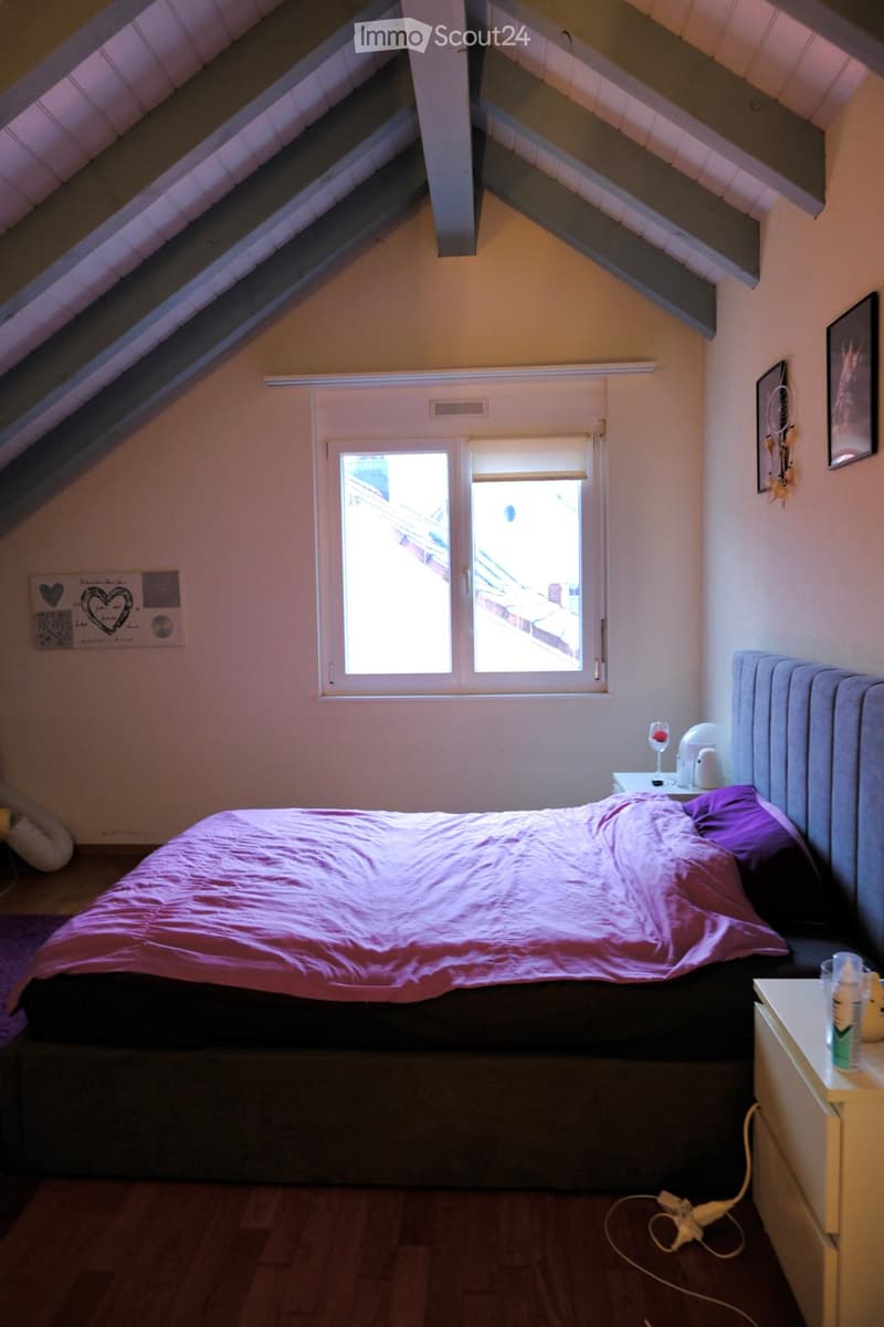 Cozy attic bedroom with slanted wooden ceiling, large window, bed with pink bedding, nightstand, lamp, and decorative items on walls.