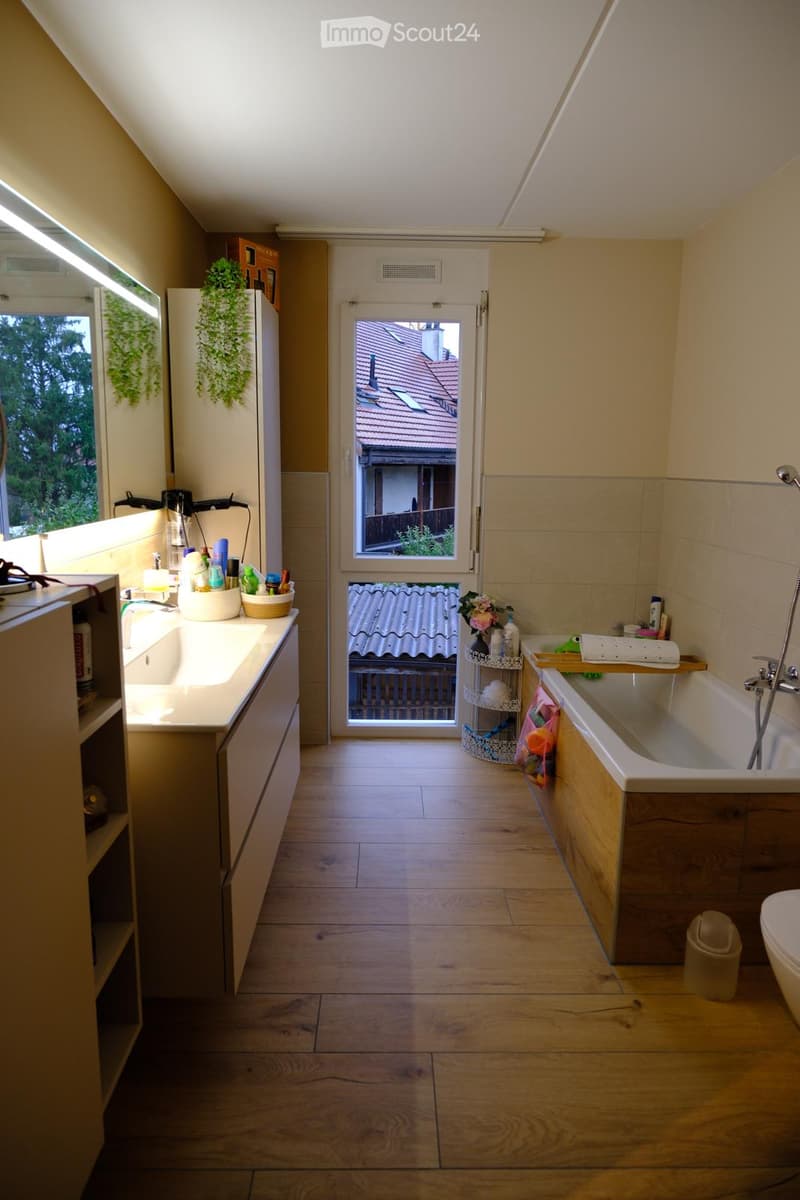 Bathroom with wooden flooring, a sink with a cabinet, a bathtub, a toilet, a shower, a mirror, a flower vase, a basket, and a towel rack. A door leads to an outdoor area with a view of a roof.