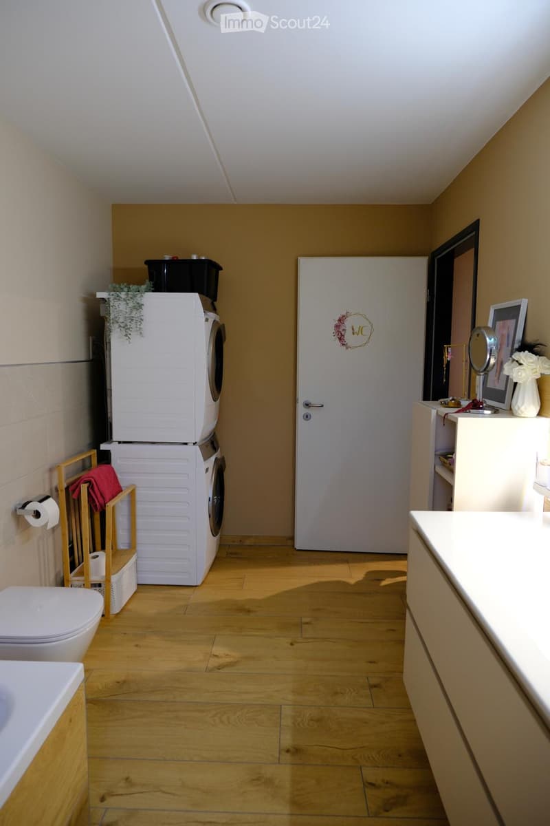 Bathroom with wooden flooring, white toilet, sink with mirror, washing machine and dryer stacked, towel rack, and decorative items.