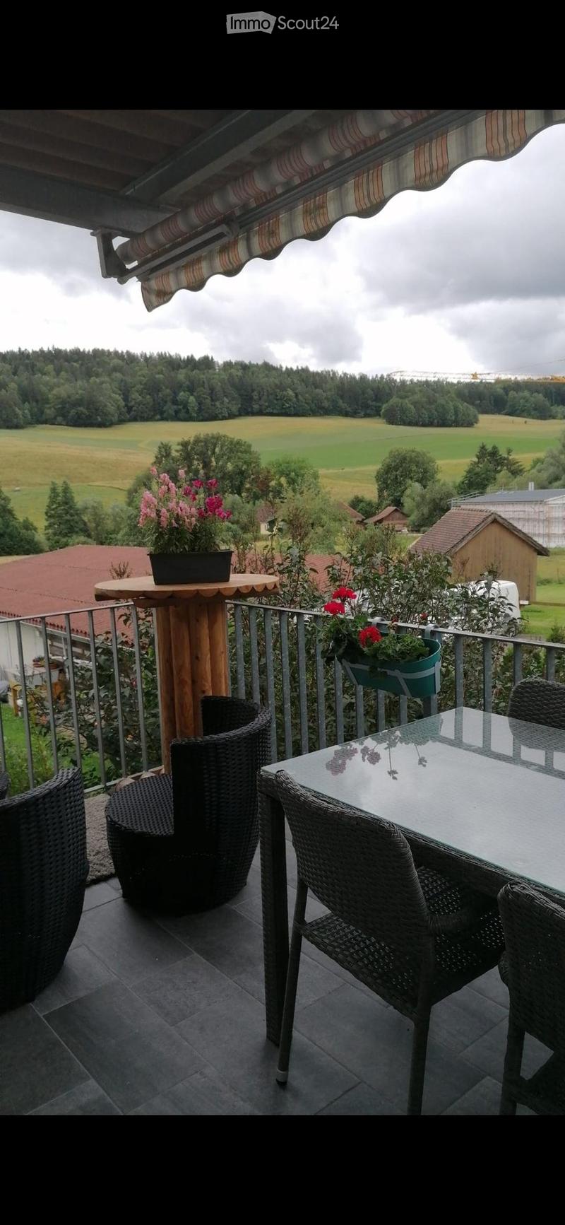 Balcony with a view of a green landscape, featuring a round wooden table, wicker chairs, and potted plants.