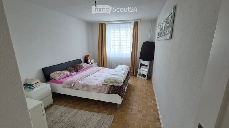 Cozy bedroom with a white bed, pink bedding, wooden nightstand, books, toys, and a small wardrobe.