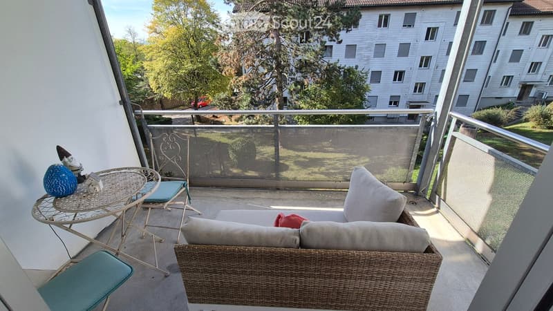 Balcony with a wicker sofa, small table, and chairs, overlooking a residential area with trees and buildings.