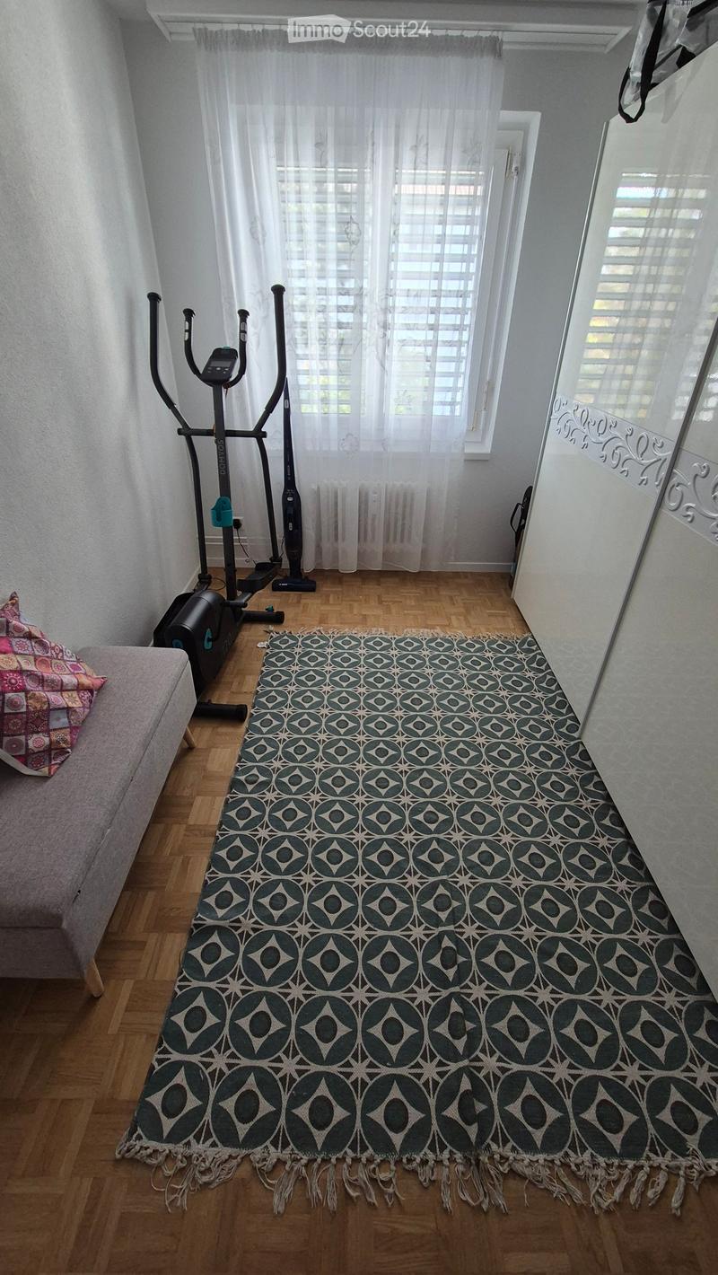 A small, well-lit room with wooden flooring, a patterned rug, a grey sofa, an exercise bike, and a large mirror on the wall.