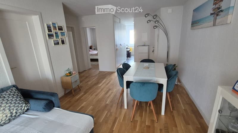 A modern, well-lit living and dining area with a sofa, coffee table, dining table with chairs, and a floor lamp. The space features wooden flooring and white walls, with a hallway leading to another room.