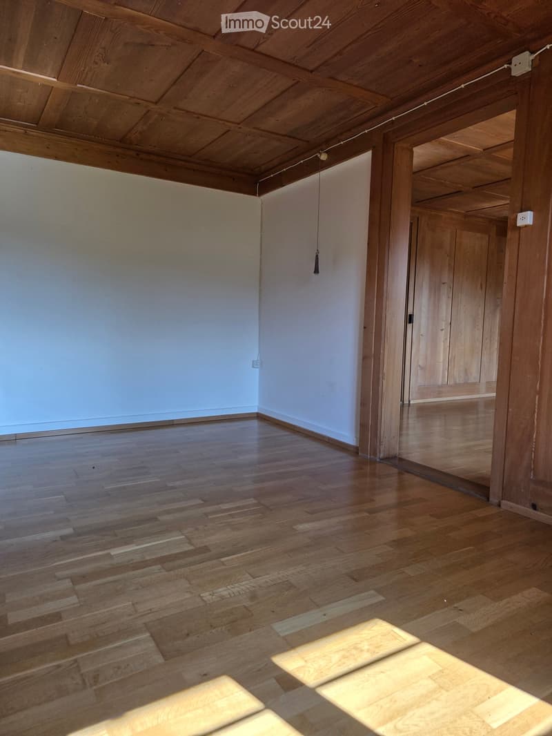 Empty room with wooden floor and exposed wooden ceiling beams, featuring a large window and a wooden door leading to another room.