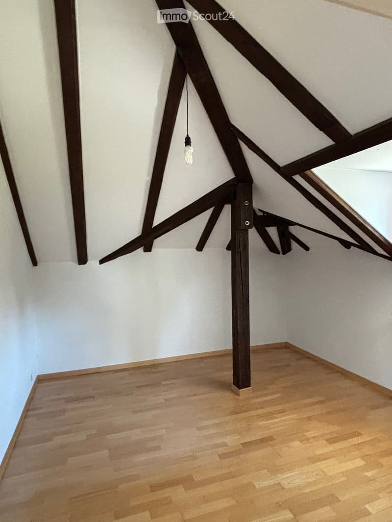 Empty attic space with wooden beams, wooden floor, and a single hanging light bulb.