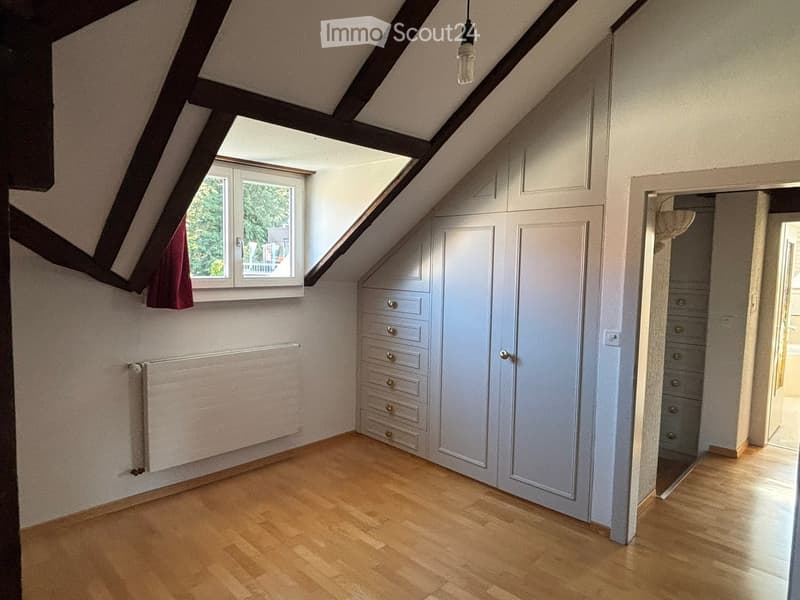 An attic room with wooden flooring, exposed wooden beams, built-in wardrobes, and a window offering a view of greenery.