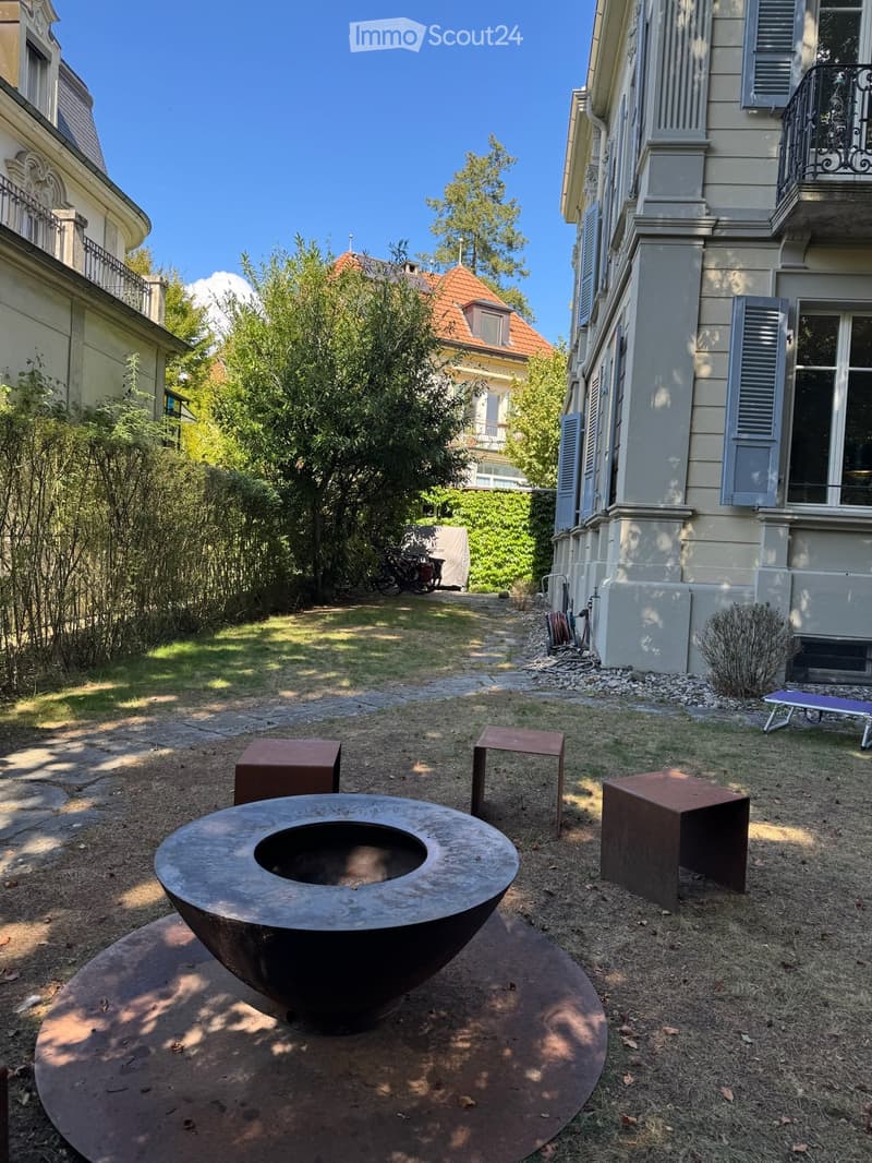 Garden area with a large metal fire bowl, four square stools, and a small lounge chair near a building with balconies and shutters.