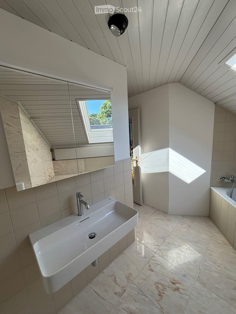 Modern bathroom with a white sink, chrome faucet, large mirror, and a bathtub. Light-colored tiles and wooden ceiling.