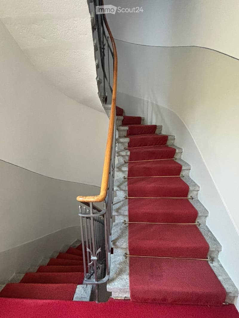 Staircase with red carpeted steps, wooden handrail, and metal balustrade.
